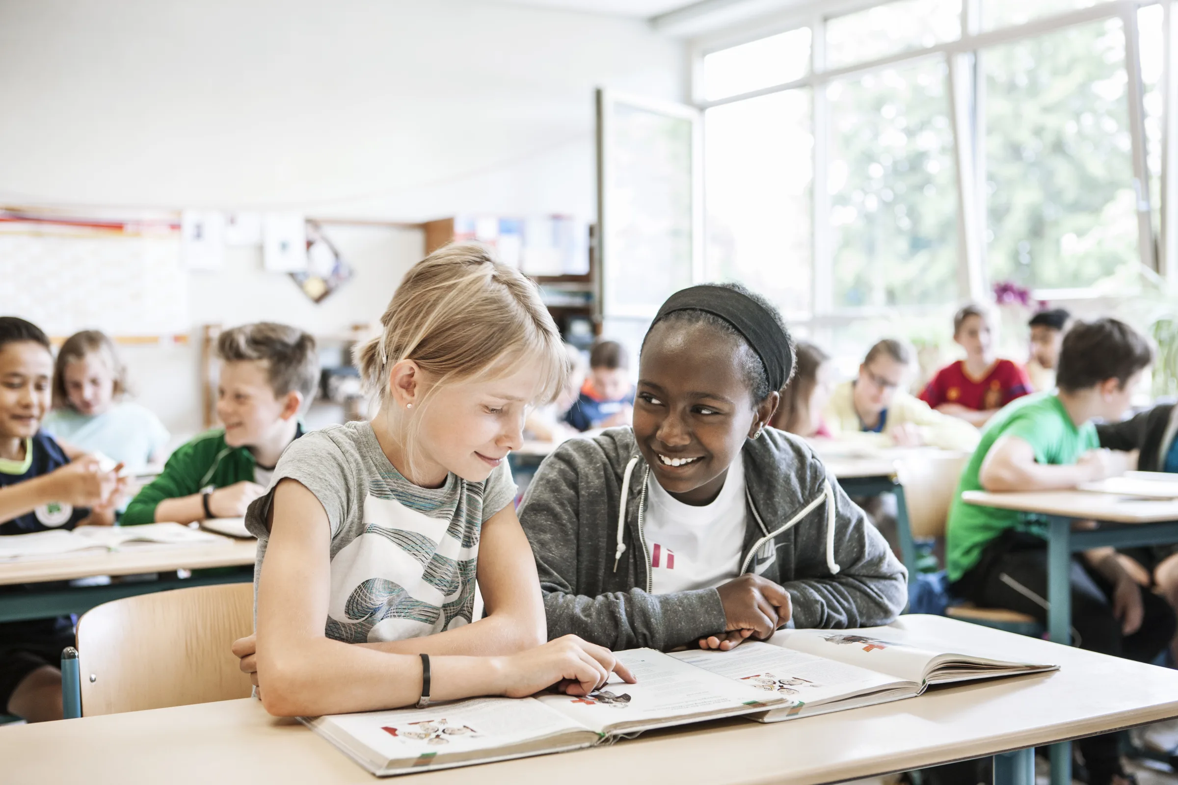 Zwei Schülerinnen lesen gemeinsam in einem Buch, dahinter arbeitet die Klasse an ihren Tischen.